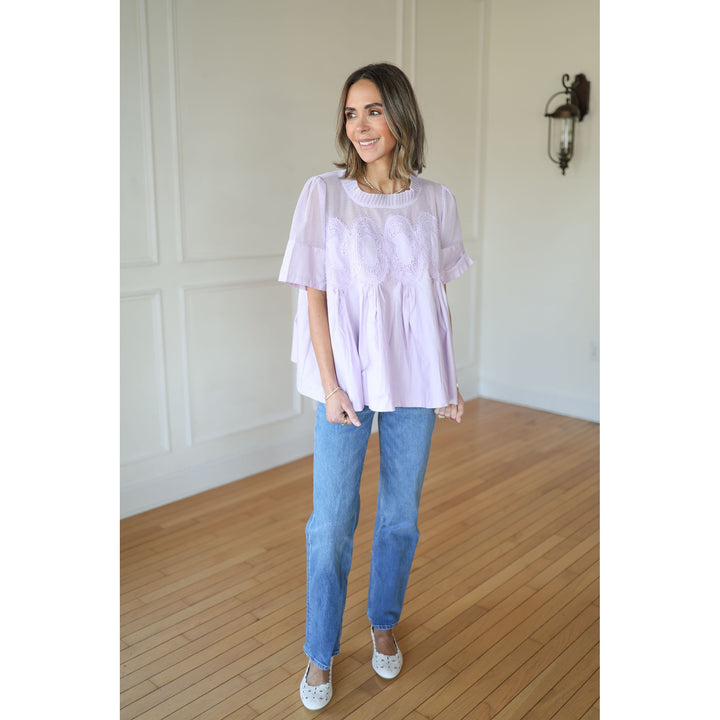 Woman wearing a light purple top and blue jeans standing in a room with wooden flooring and white walls.