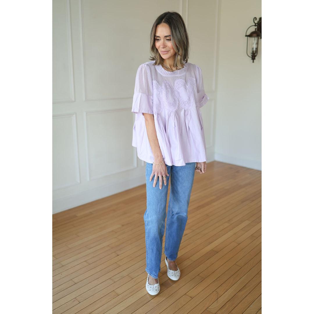 Woman wearing a light purple blouse and blue jeans standing in a room with wooden flooring and white walls.