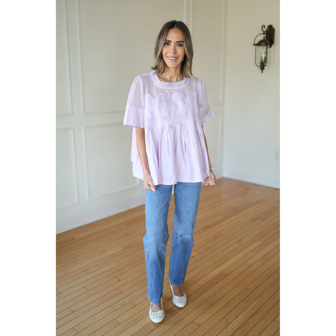 Woman wearing a light purple blouse and blue jeans standing in a room with wooden flooring and white walls.