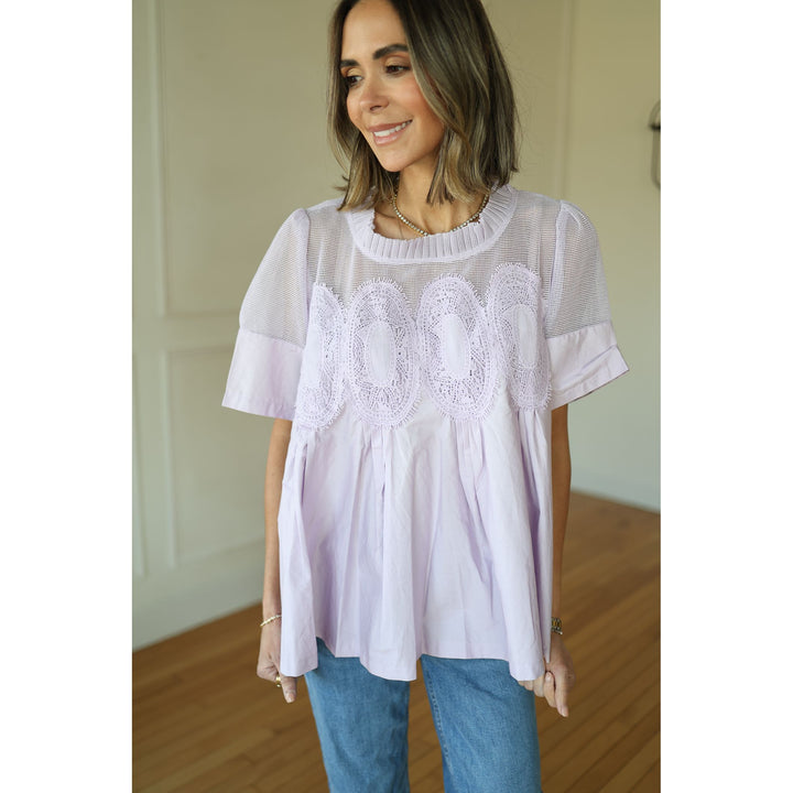 Woman wearing a light purple lace top and blue jeans indoors.