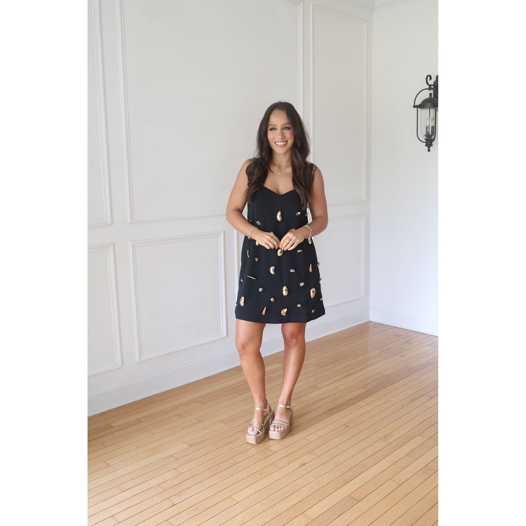 Woman in a black dress with gold patterns standing in a room with white walls and wooden floor.
