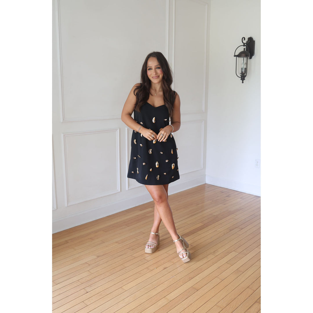 Woman in a black dress with gold patterns standing in a room with wooden flooring and white walls.