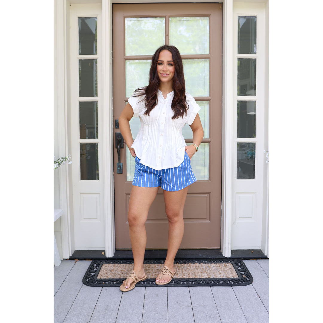 Woman standing in front of a door wearing a white blouse and blue striped shorts.