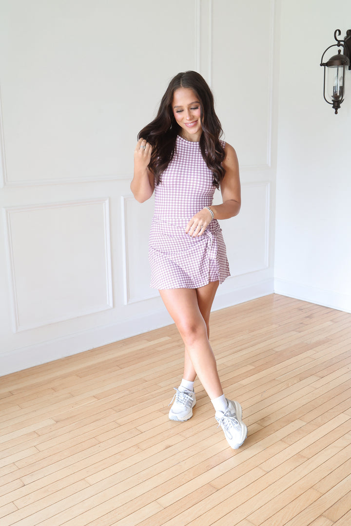 A woman modeling a sleeveless, high-neck mini dress with a pink and white gingham pattern, paired with white sneakers