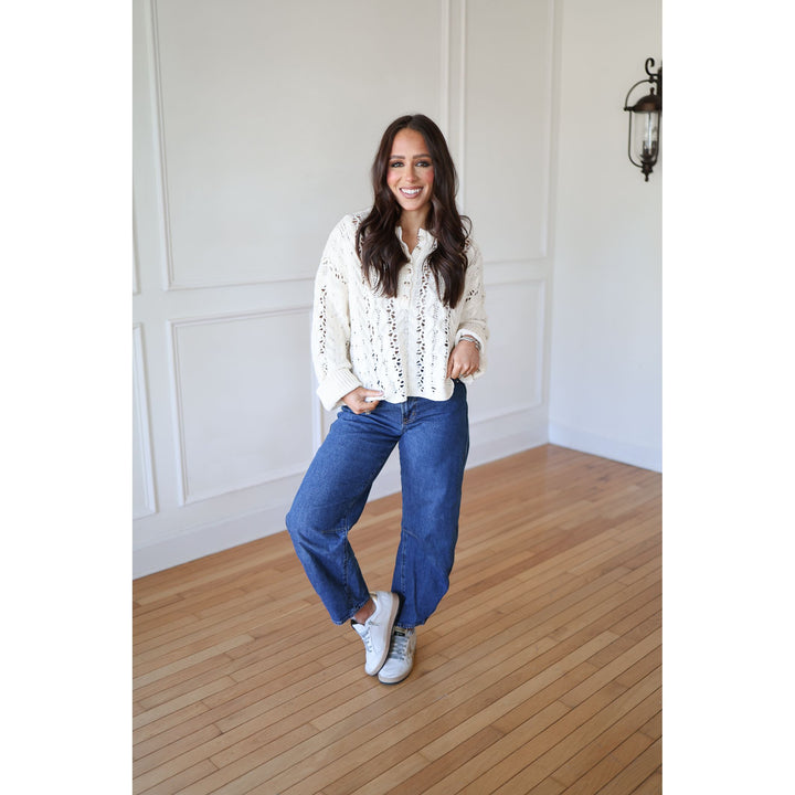 Woman wearing a white blouse with lace details and blue jeans standing on a wooden floor.