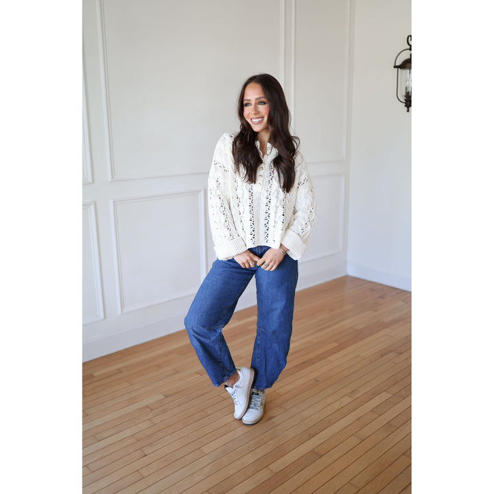 Woman wearing a white blouse with lace details and blue jeans standing in a room with wooden flooring and white walls.