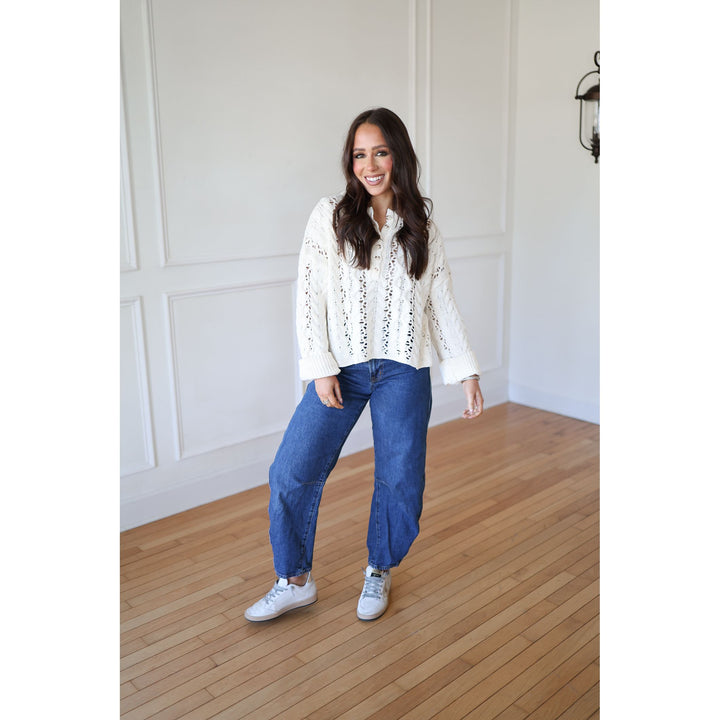 Woman wearing a white blouse with lace details and blue jeans standing in a room with wooden flooring and white walls.