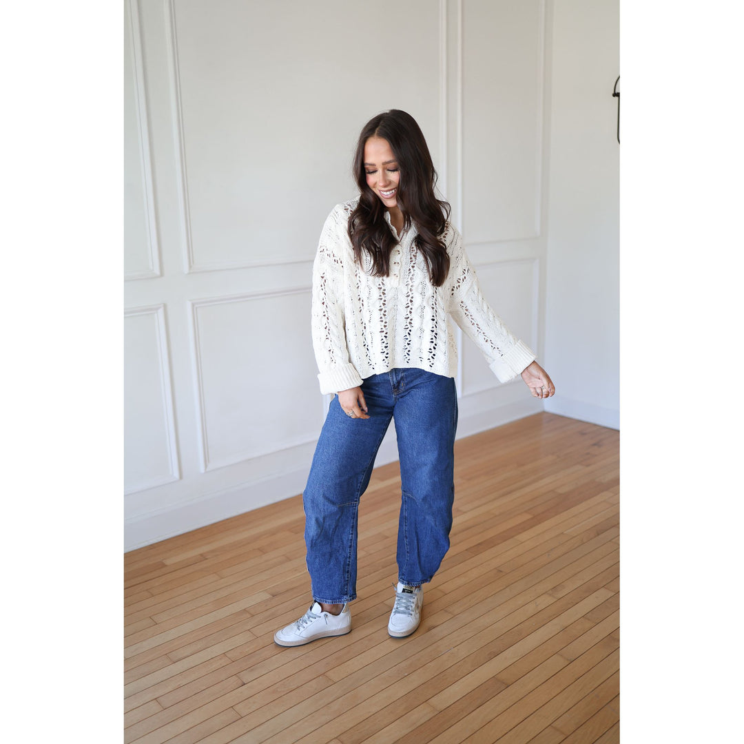 Woman wearing a white lace blouse and blue jeans standing on a wooden floor.