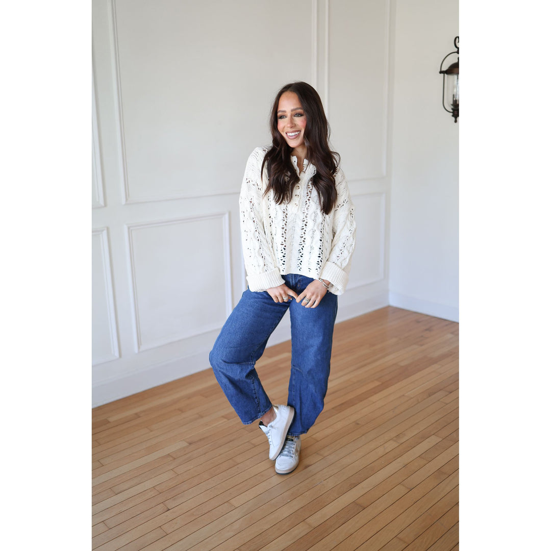 Woman wearing a white blouse with black patterns and blue jeans standing on a wooden floor.