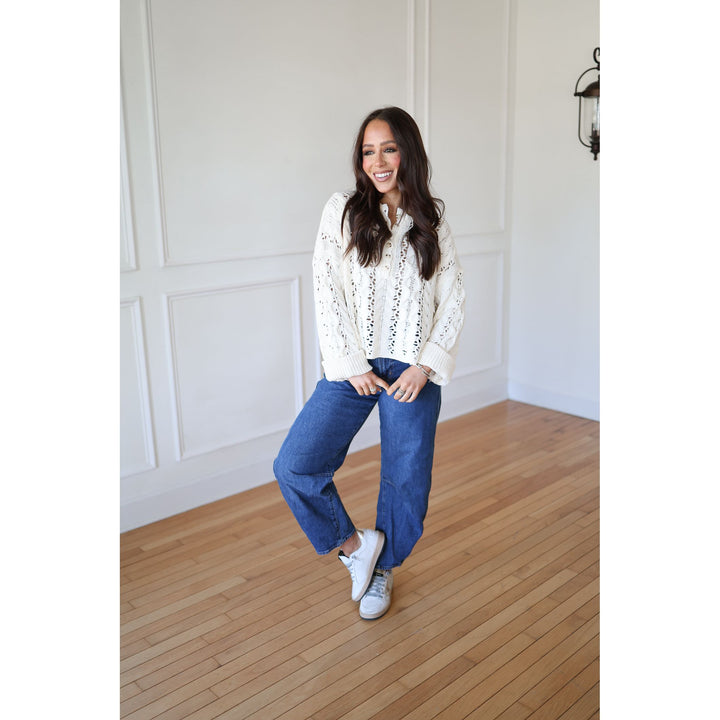 Woman wearing a white blouse with black patterns and blue jeans standing on a wooden floor.