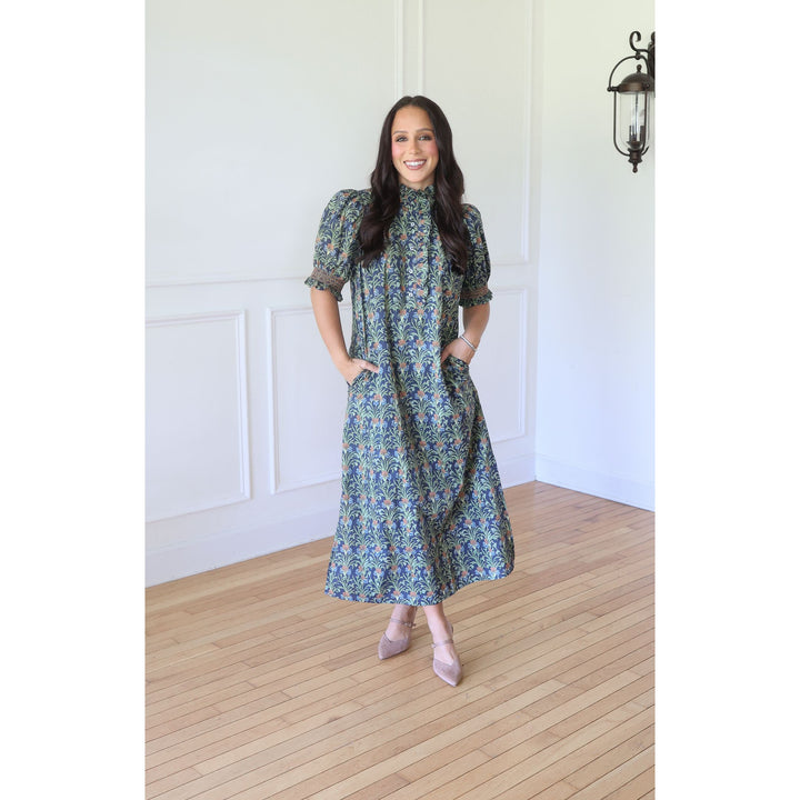 A woman stands in a bright room with white paneled walls and light wood floors, wearing a long-sleeve midi dress with a blue and green floral pattern, paired with beige pointed-toe heels.
