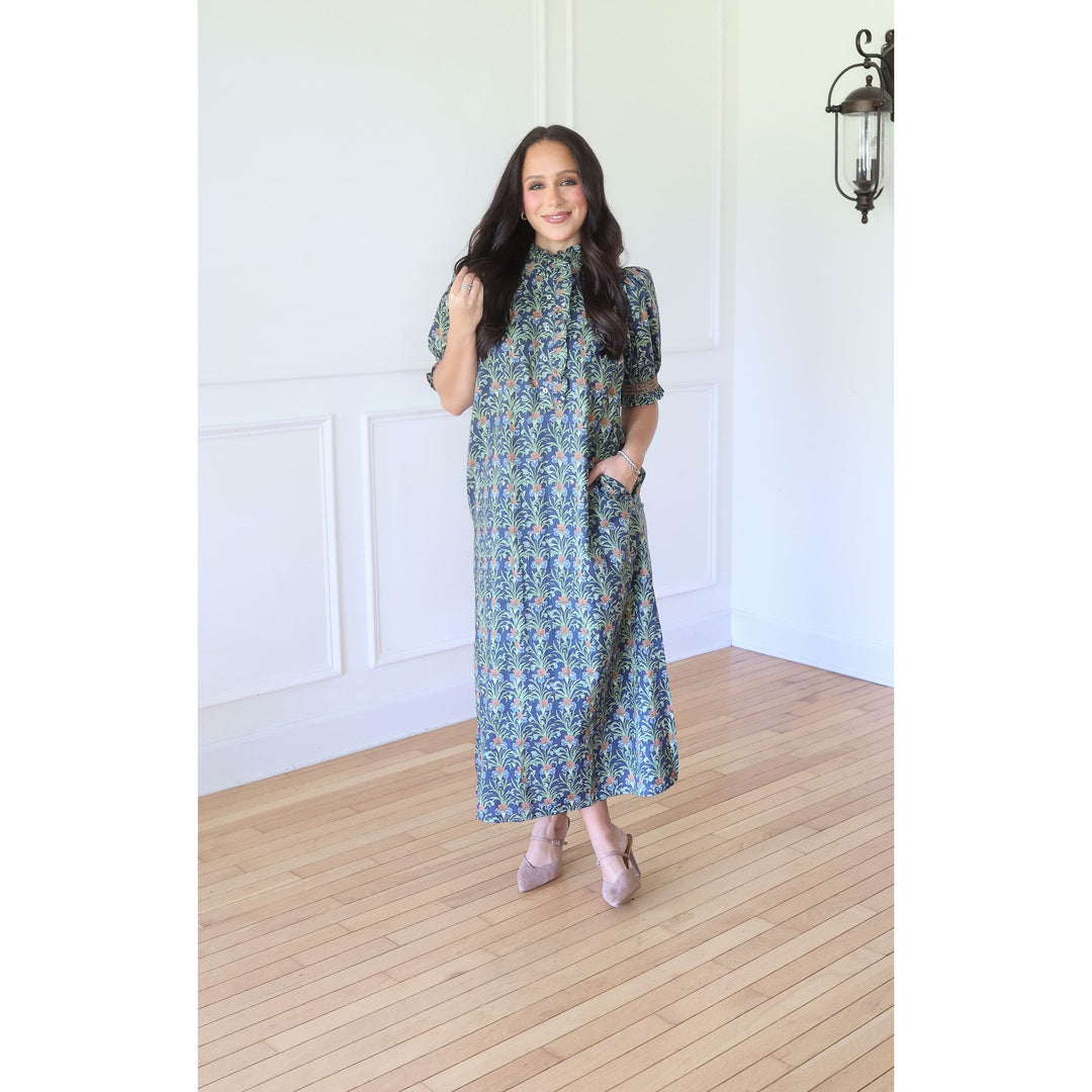 A woman modeling a blue and green patterned midi dress with short sleeves, a high collar, and side pockets, standing in a bright room with white walls and light wood floors.