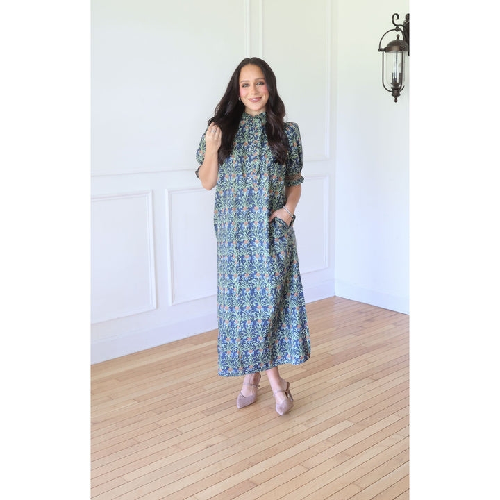 A woman modeling a blue and green patterned midi dress with short sleeves, a high collar, and side pockets, standing in a bright room with white walls and light wood floors.