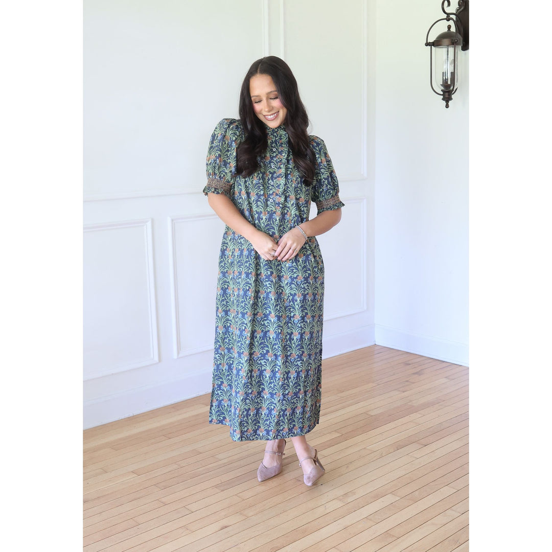 A woman wearing a short-sleeved midi dress with a blue and green paisley pattern, standing in a bright room with white walls and light wood floors.