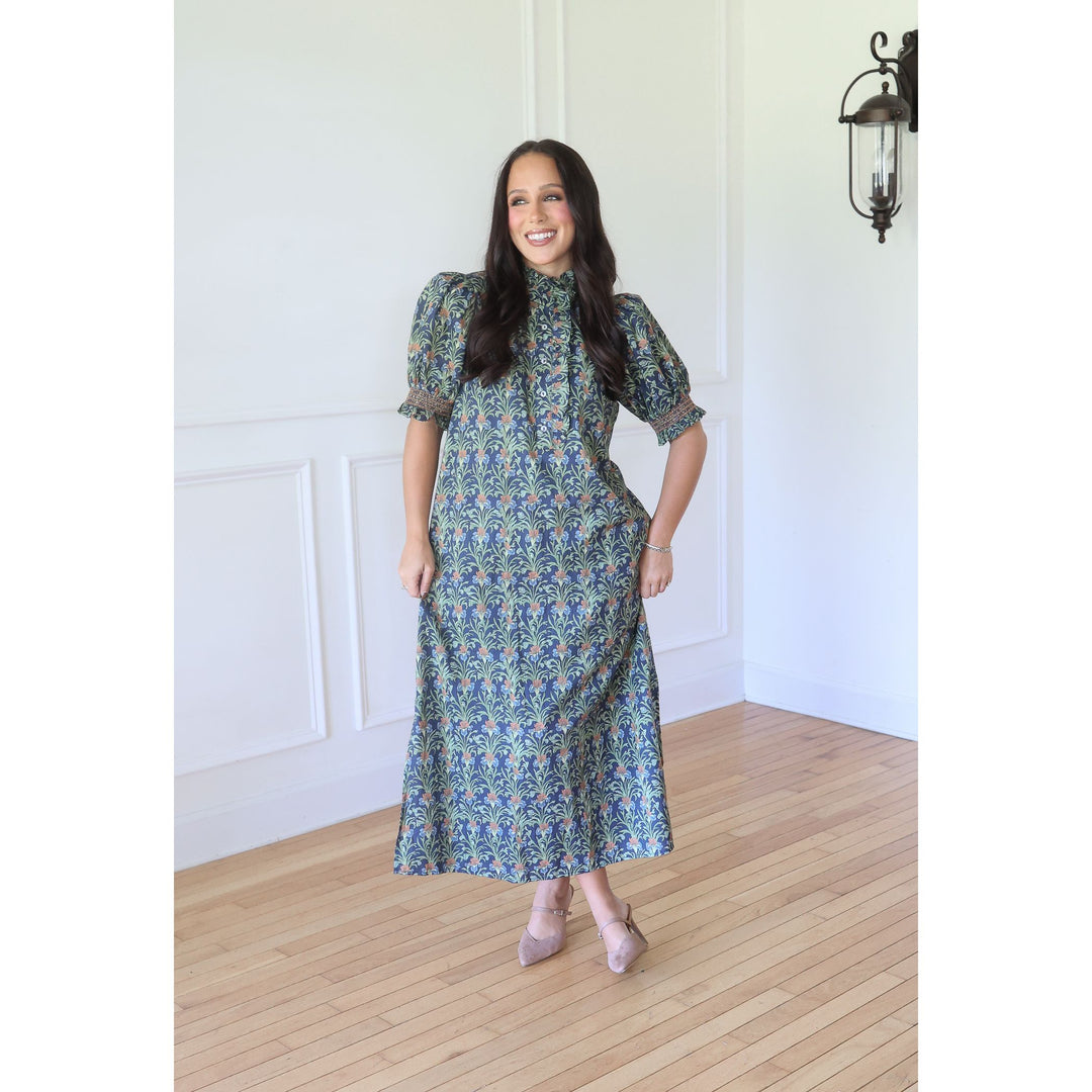 A woman modeling a short-sleeved midi dress with a blue and green floral pattern, standing in a bright room with white walls and light wood floors.