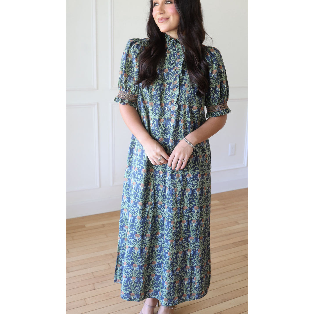 A woman wearing a long-sleeved midi dress with a blue, green, and orange botanical pattern, standing in a room with light wood floors and white paneled walls.