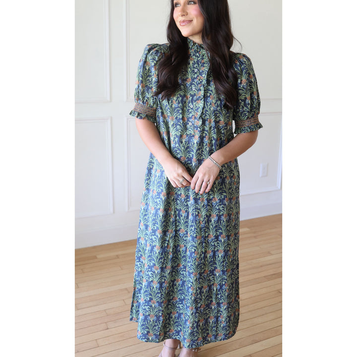 A woman wearing a long-sleeved midi dress with a blue, green, and orange botanical pattern, standing in a room with light wood floors and white paneled walls.