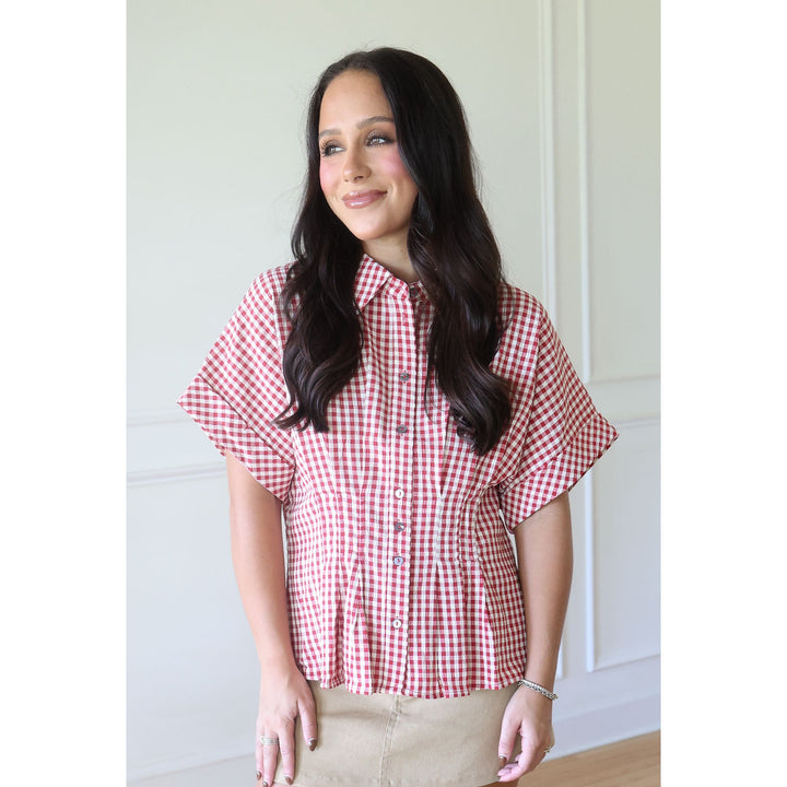 A woman with long dark hair wearing a red and white gingham short-sleeve button-up shirt and beige pants, standing in a brightly lit indoor setting with white paneled walls.