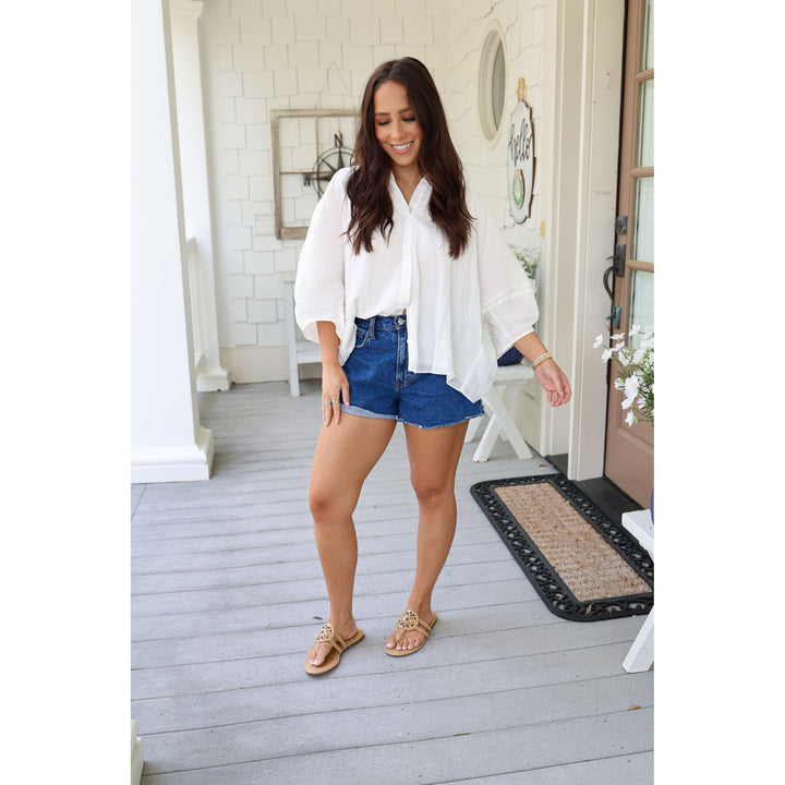 Woman in white blouse and blue shorts standing on a wooden deck.