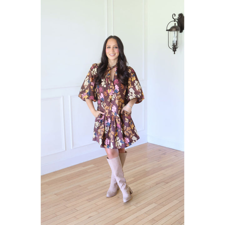 A woman modeling a short, loose floral dress with wide sleeves and a tie waist, paired with knee-high beige boots, standing in a bright room with white paneled walls and light wood floors.