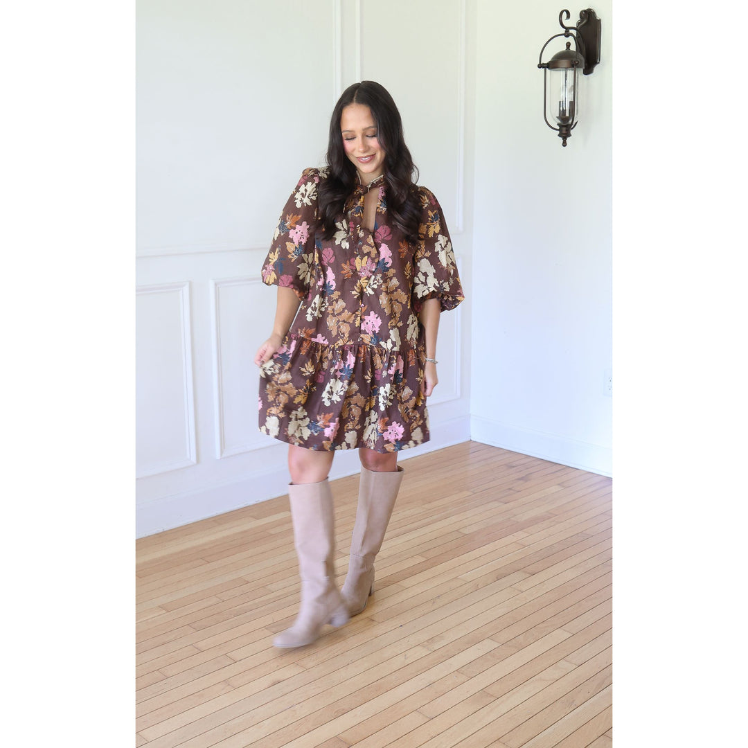 A woman wearing a brown floral tiered mini dress with puff sleeves and beige knee-high boots, standing in a room with white paneled walls and light wood floors.