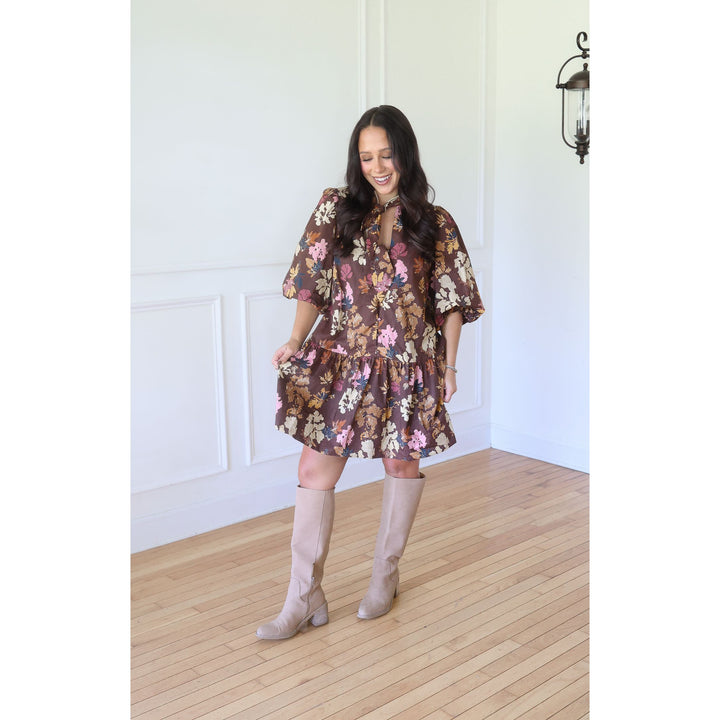 A woman modeling a brown floral tiered mini dress with puff sleeves, paired with beige knee-high boots, standing in a bright room with white paneled walls and a light wood floor.