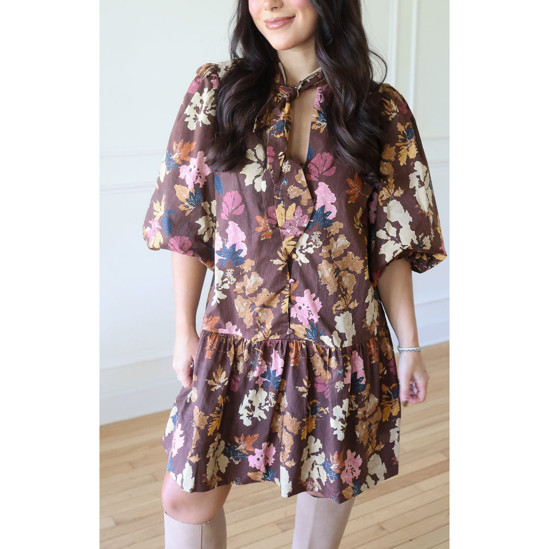 A woman wearing a brown short-sleeve mini dress with a colorful floral print in pink, beige, and blue, paired with beige knee-high socks, standing in a brightly lit room with light wood floors.