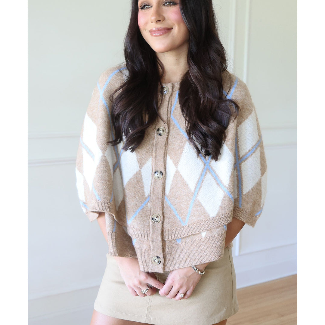 A woman wearing a beige and white argyle knit cardigan with light blue accents, paired with a beige mini skirt, standing in a brightly lit indoor setting.