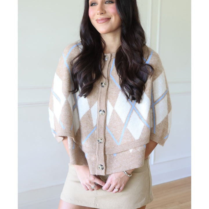 A woman wearing a beige and white argyle knit cardigan with light blue accents, paired with a beige mini skirt, standing in a brightly lit indoor setting.