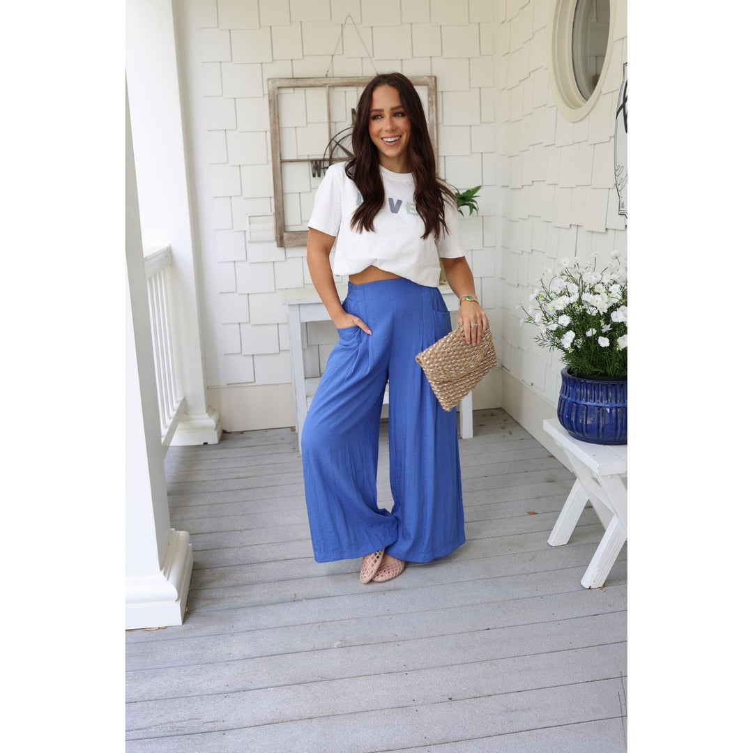 Woman in blue pants and white top standing on a wooden deck with a white wall and decorative elements in the background.