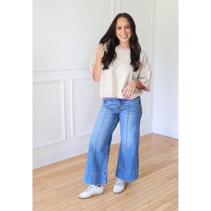A woman modeling a cream knit sweater with pink trim, blue wide-leg jeans, and white sneakers in a bright room with white paneled walls and light wood floors.