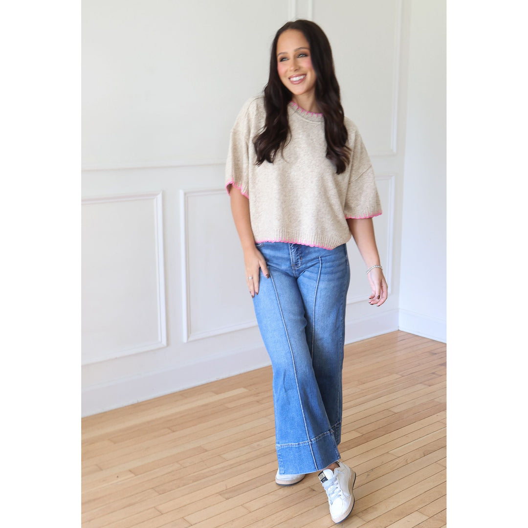 A woman modeling a beige knit short-sleeve sweater with pink trim, paired with blue jeans and white sneakers, standing in a bright room with white paneled walls and light wood floors.