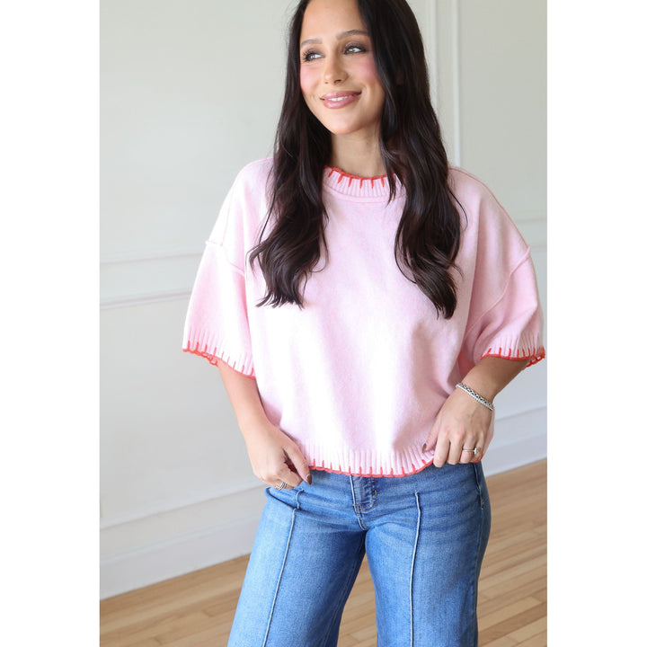 A woman wearing a light pink knit sweater with contrasting coral-red scalloped trim on the crew neck and short sleeves, paired with blue jeans, standing in a brightly lit room with white walls and light wood floors.