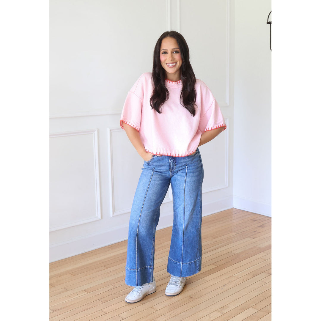 A woman stands in a bright room with white paneled walls and light wood floors, wearing a light pink short-sleeve top with scalloped edges, blue wide-leg jeans, and white sneakers.