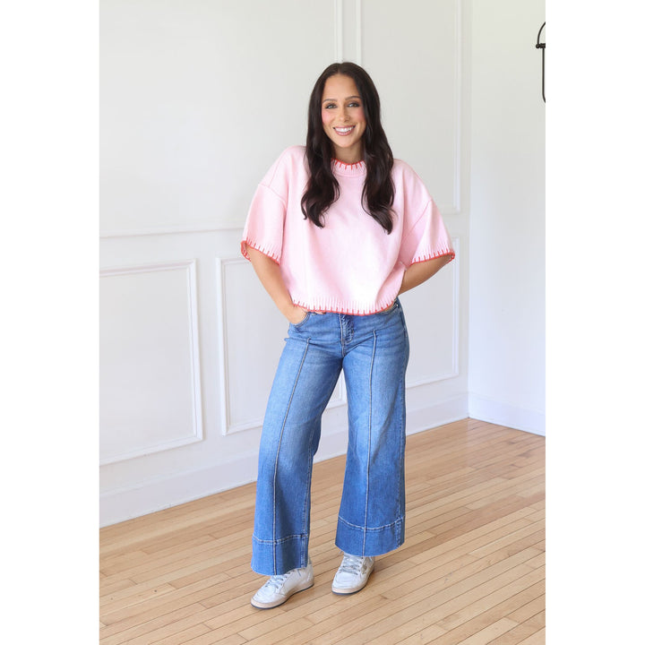 A woman stands in a bright room with white paneled walls and light wood floors, wearing a light pink short-sleeve top with scalloped edges, blue wide-leg jeans, and white sneakers.