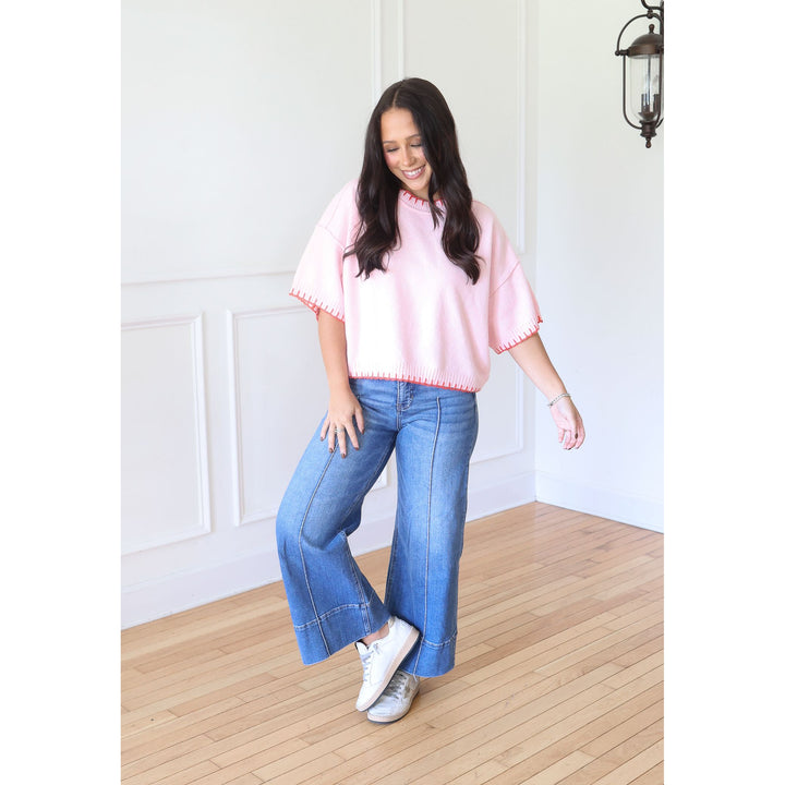 A woman modeling a light pink short-sleeve top with red trim and blue wide-leg jeans, standing on a light wood floor against a white paneled wall.