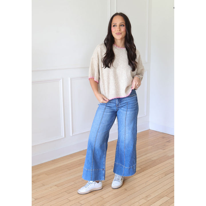 A woman modeling a beige knit short-sleeve sweater with pink trim and wide-leg blue jeans, standing on a light wood floor against a white paneled wall.
