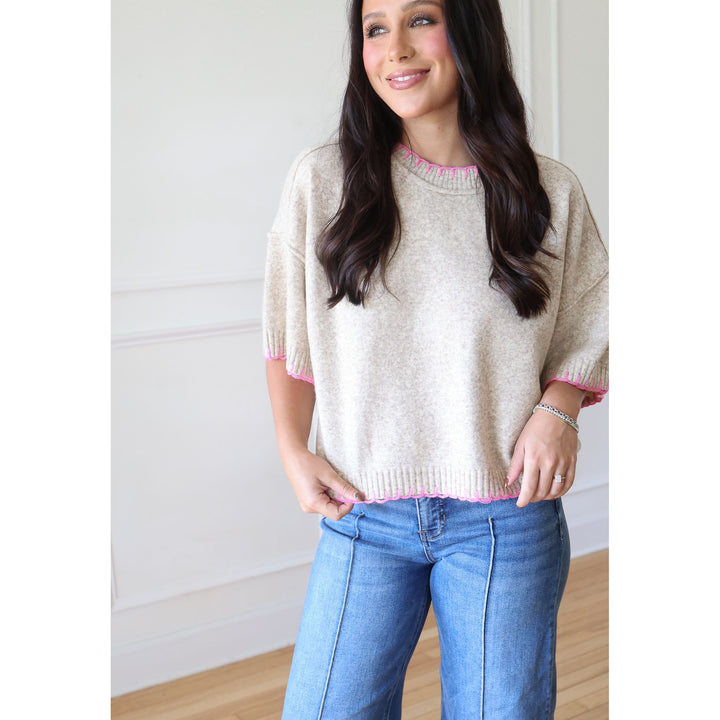 A woman wearing a cream-colored knit sweater with pink trim on the collar and cuffs, paired with blue jeans, standing in a brightly lit room with white walls and light wood flooring.