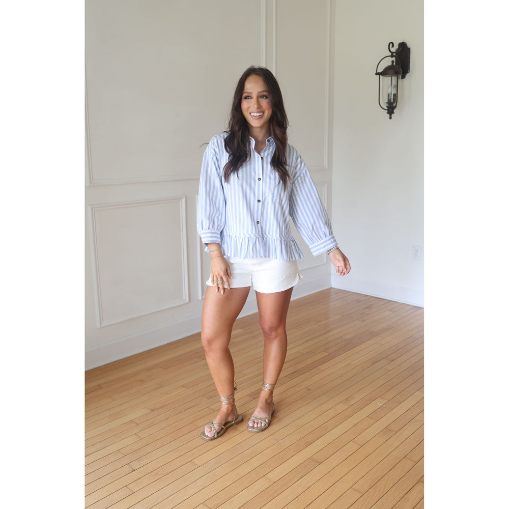 Woman wearing a striped shirt and white shorts standing in a room with wooden flooring and white walls.