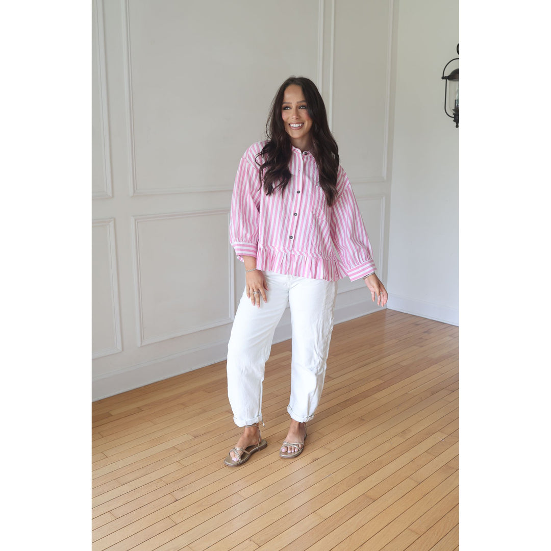 Woman wearing a pink striped shirt and white pants standing in a room with wooden flooring and white walls.