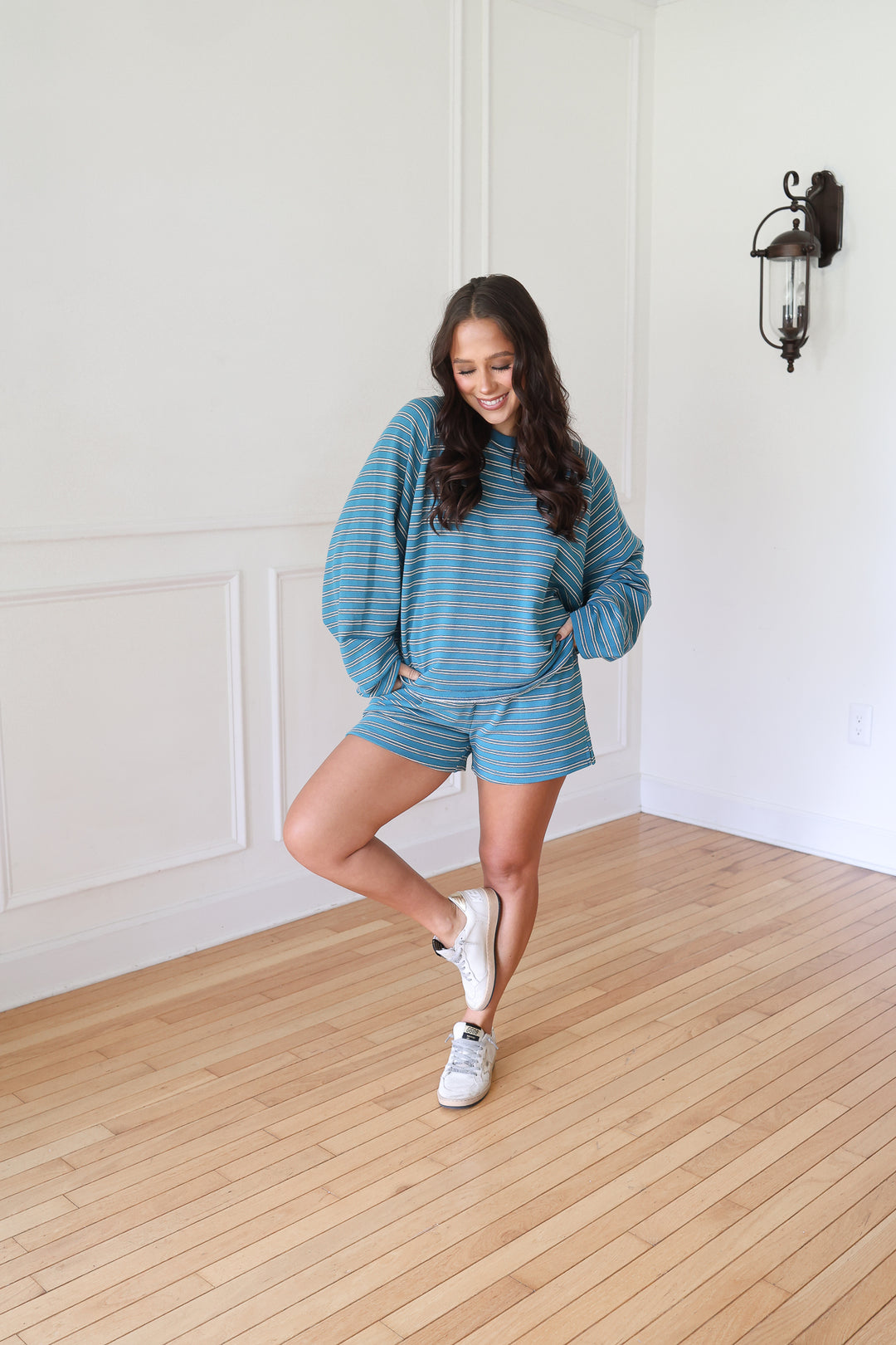 A woman posing in a matching blue and white striped lounge set, consisting of a long-sleeve sweatshirt and shorts, paired with white sneakers