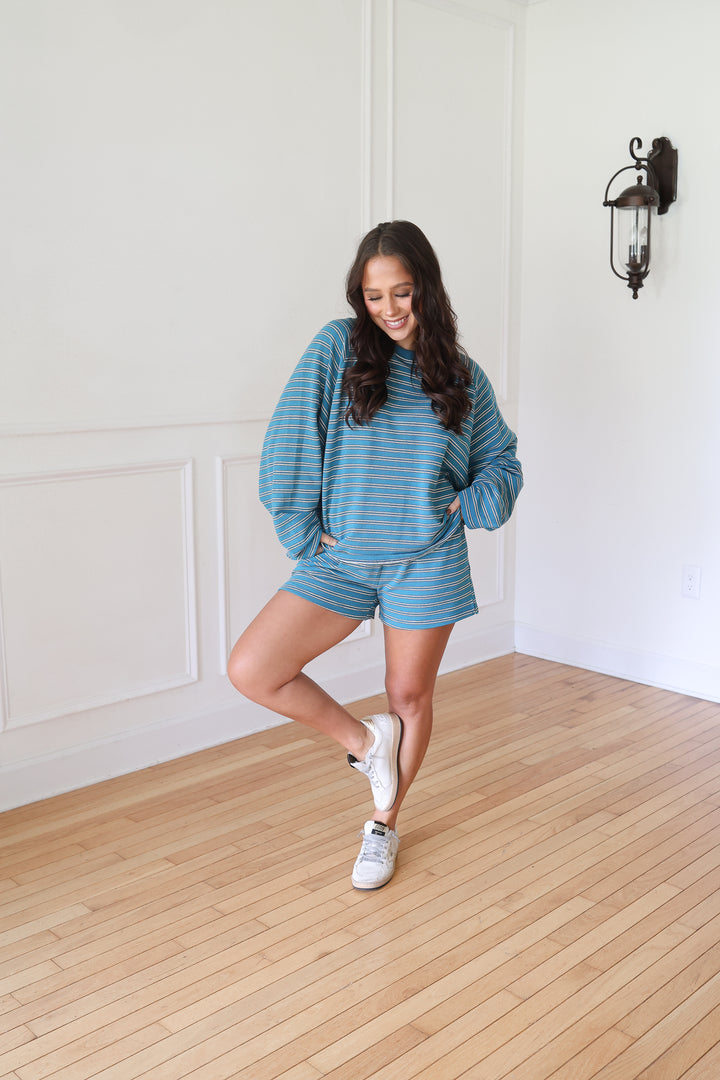 A woman posing in a matching blue and white striped lounge set, consisting of a long-sleeve sweatshirt and shorts, paired with white sneakers