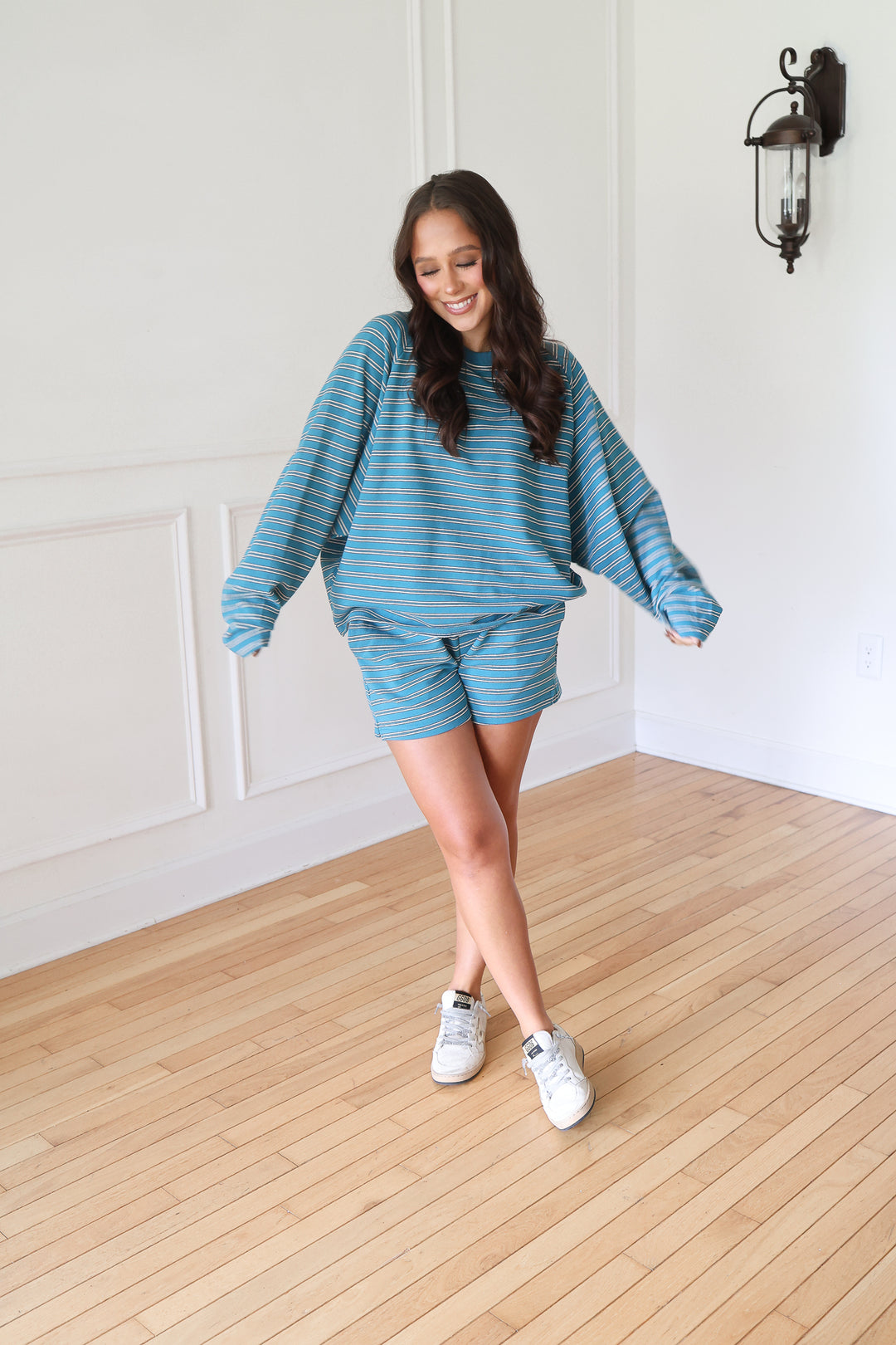 A woman modeling a matching blue and white striped lounge set consisting of a long-sleeve sweatshirt and shorts, paired with white sneakers