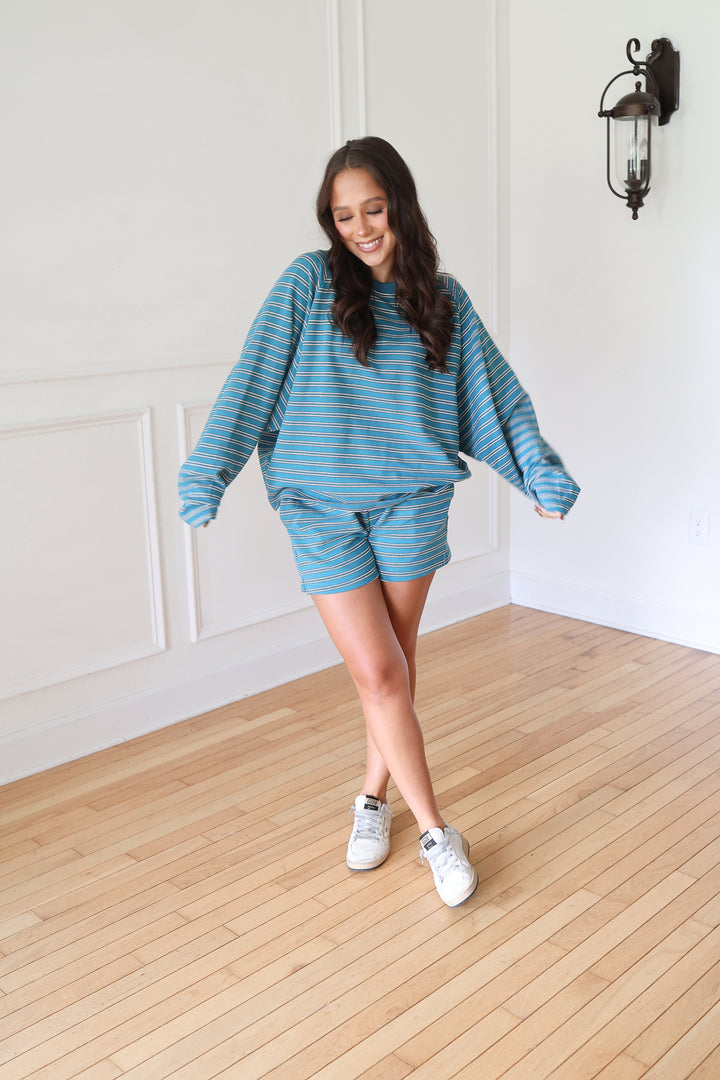 A woman modeling a matching blue and white striped lounge set consisting of a long-sleeve sweatshirt and shorts, paired with white sneakers