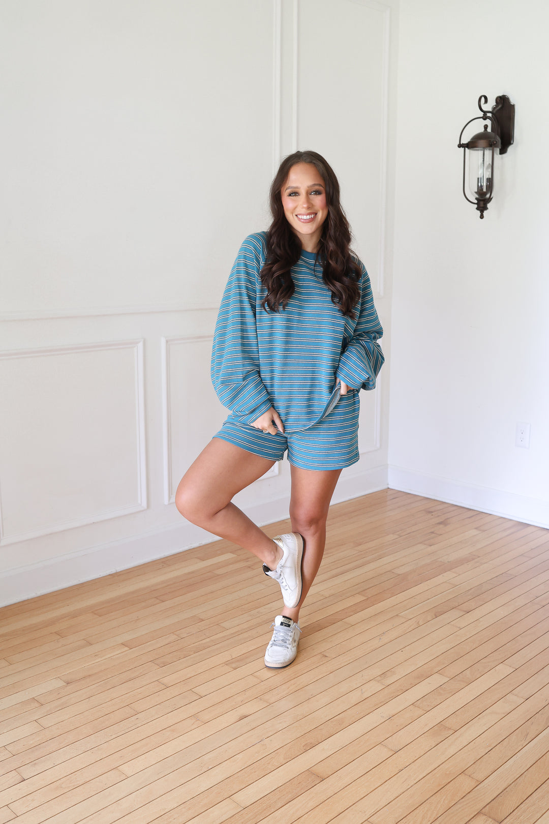 A woman posing in a matching blue and gray striped lounge set consisting of a long-sleeve sweatshirt and shorts, paired with white sneakers 