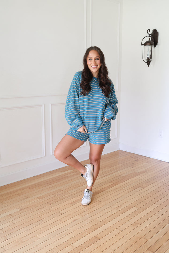 A woman posing in a matching blue and gray striped lounge set consisting of a long-sleeve sweatshirt and shorts, paired with white sneakers 