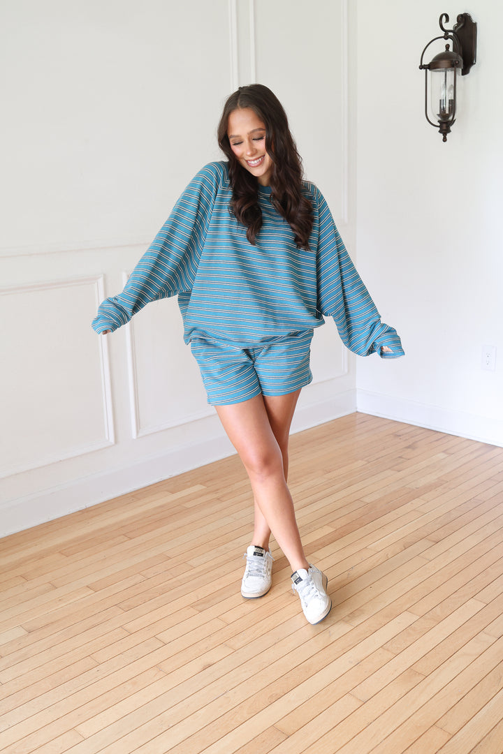 A woman modeling a matching lounge set consisting of a blue and white striped long-sleeve top and shorts, paired with white sneakers