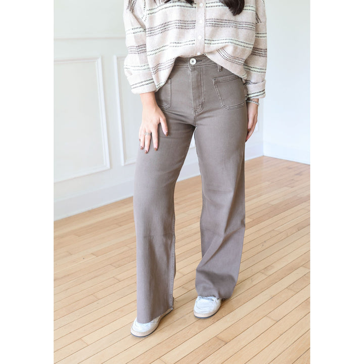 A person wearing high-waisted, wide-leg taupe gray pants, a beige striped button-up shirt, and white sneakers, standing on a light wood floor against a white paneled wall.