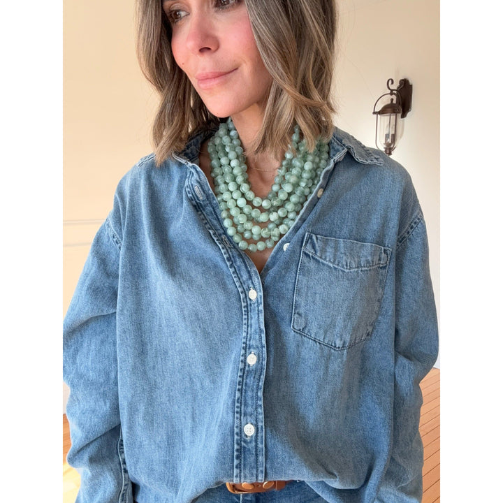 Person wearing a blue denim shirt and a long green beaded necklace indoors.