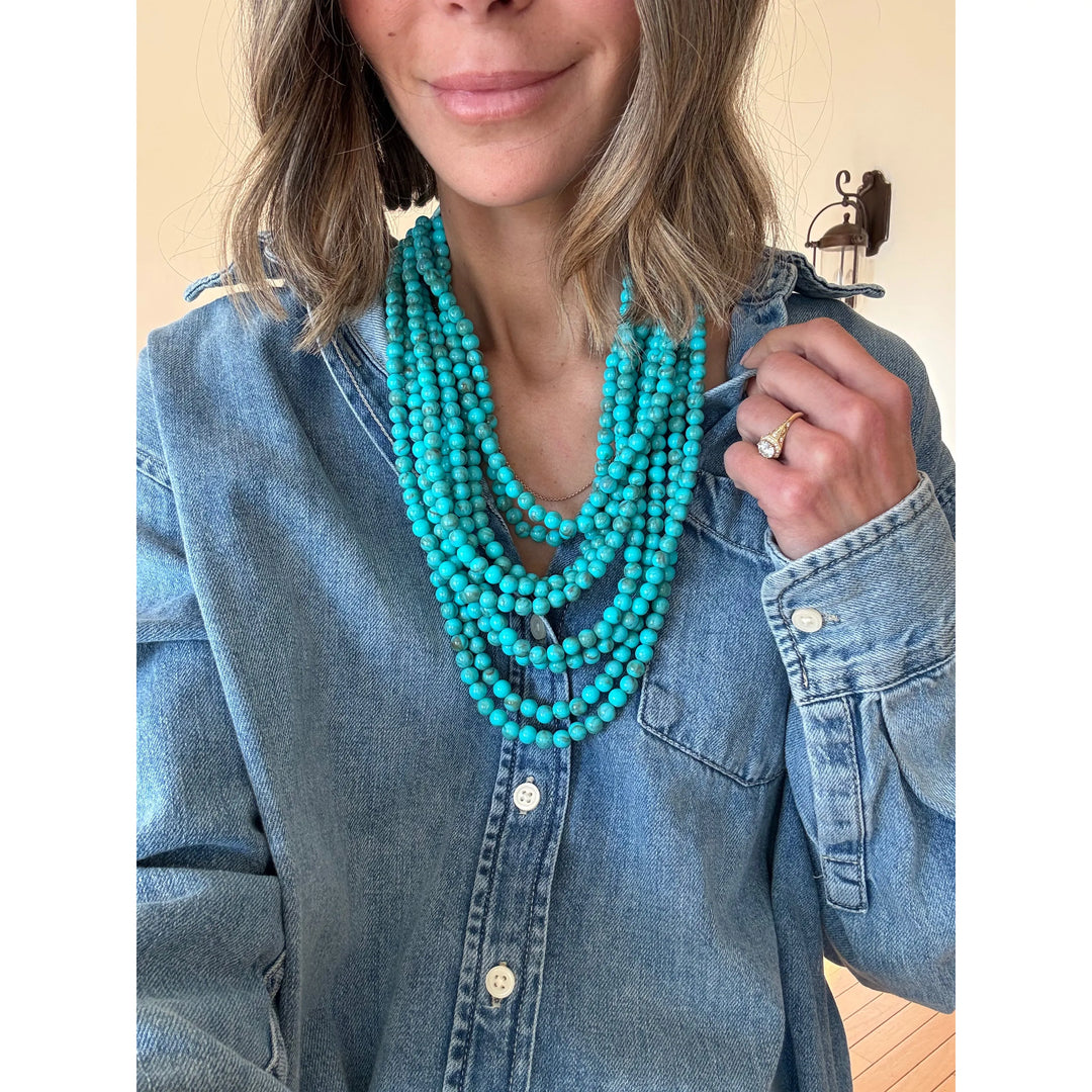 Woman wearing a blue denim shirt and turquoise beaded necklace indoors.